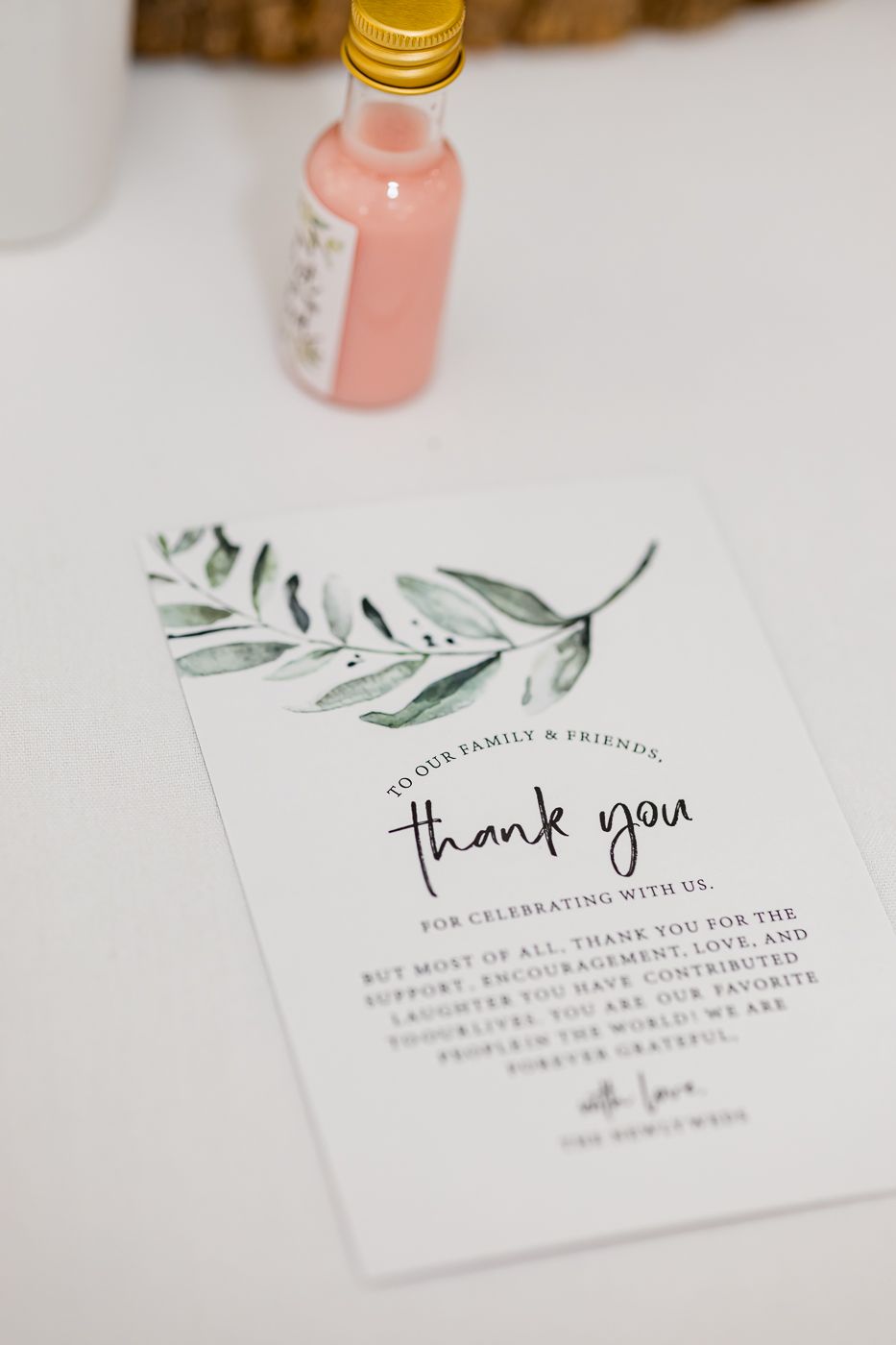 Elegant wedding thank you card with watercolor greenery beside a pink mini bottle favor on white table.