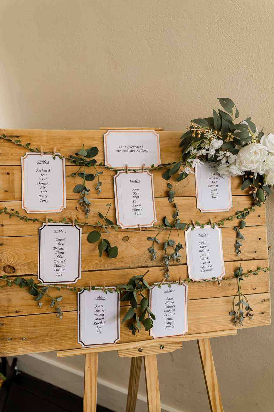 Rustic wooden easel wedding seating chart with greenery garland and white floral arrangement displaying table assignments on cards.