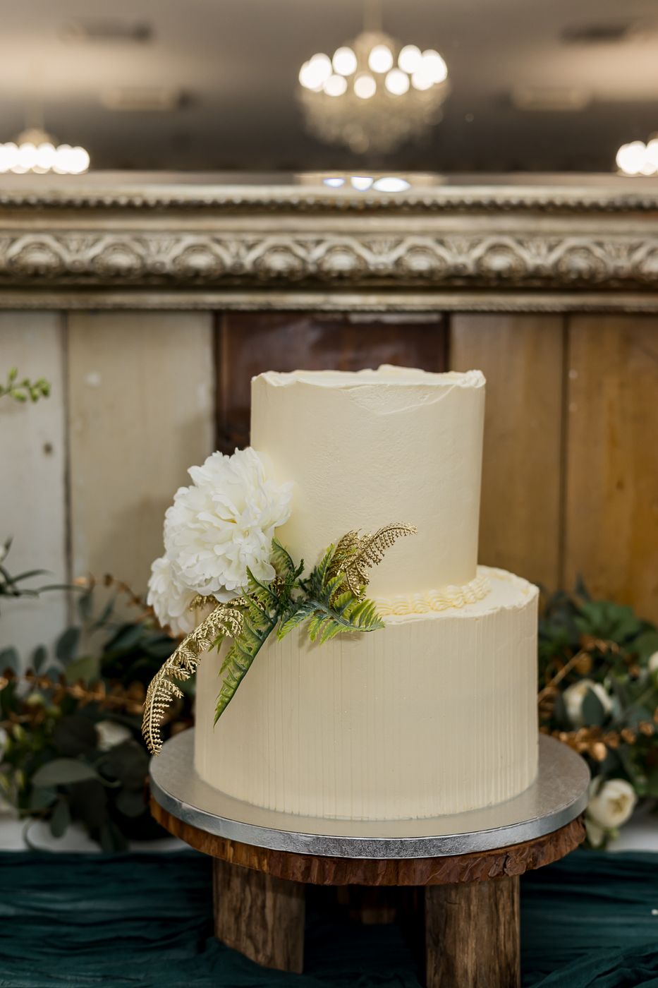 Two-tier white wedding cake with white peony and fern decor on rustic wood stand.