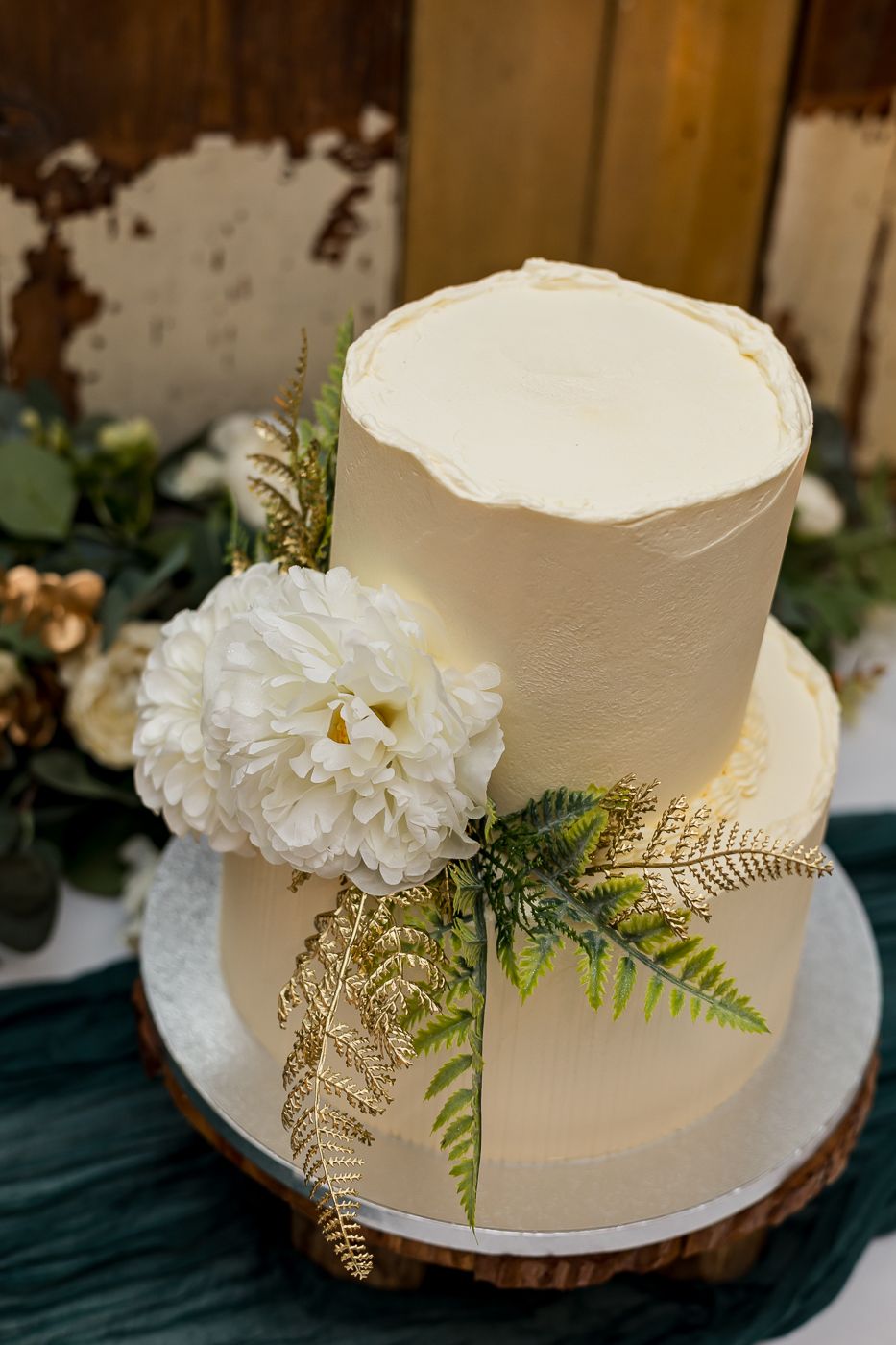 Two-tier white wedding cake with white peony and gold fern accents on a rustic wood slice stand.