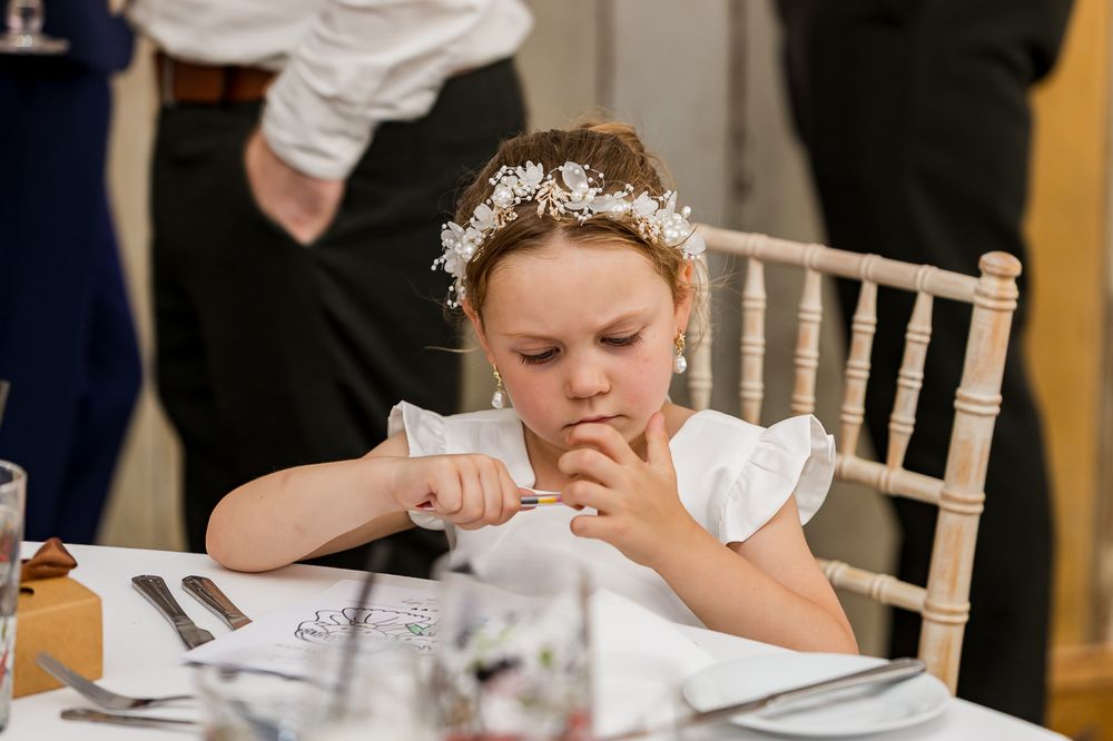 Young flower girl in white dress and floral headpiece sits at wedding reception table, looking down thoughtfully.