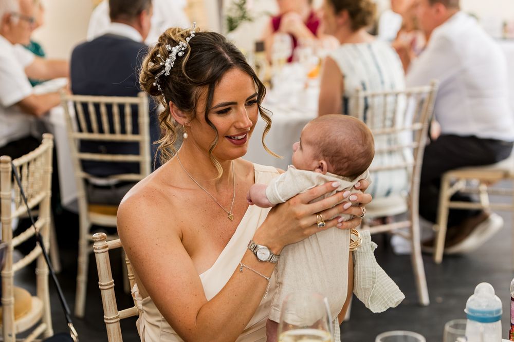 Bride in white dress holding a newborn baby at a wedding reception, smiling warmly in an elegant venue.