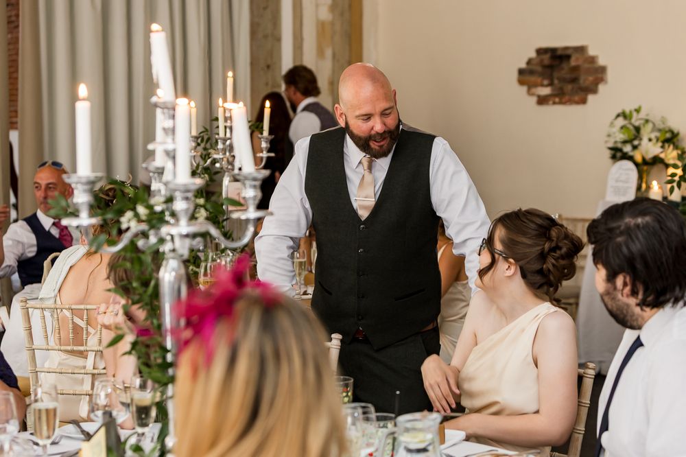 A bald groom in a vest chats with the bride seated at a decorated wedding reception table with candelabras.
