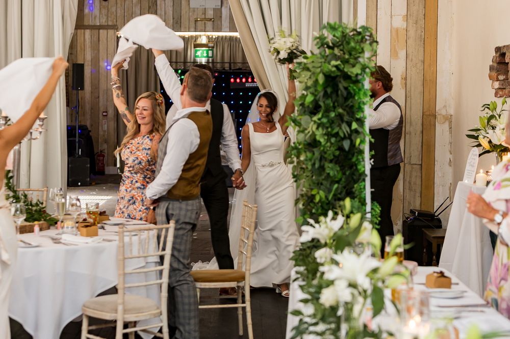 Bride and groom celebrate wedding reception entrance in decorated barn venue with guests cheering.