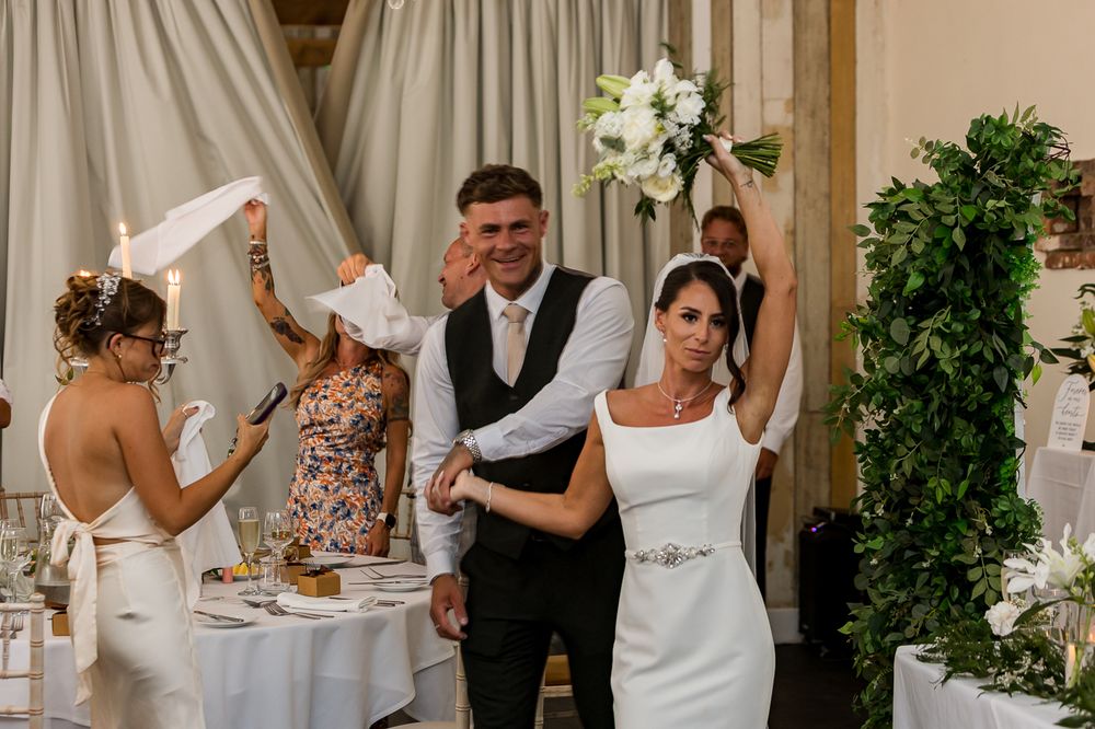 Bride raises bouquet while groom smiles during wedding reception celebration indoors with guests and greenery decor.