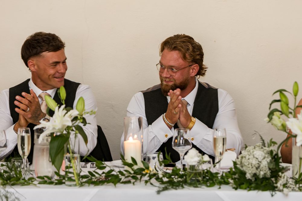 Two grooms clapping and smiling at wedding reception table decorated with white flowers and candles.