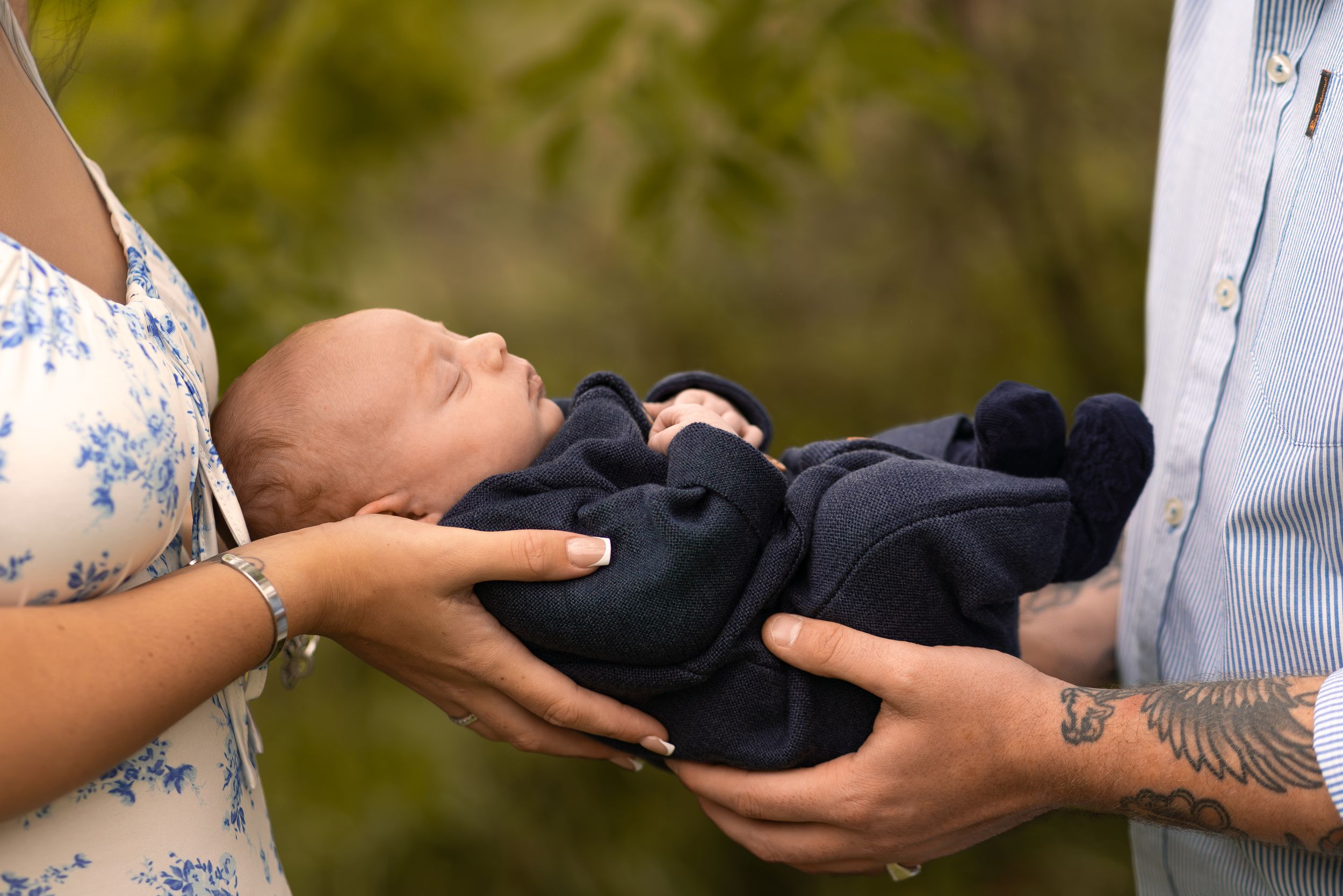 Parents cradling a sleeping newborn baby dressed in a navy blue outfit outdoors with green foliage background.