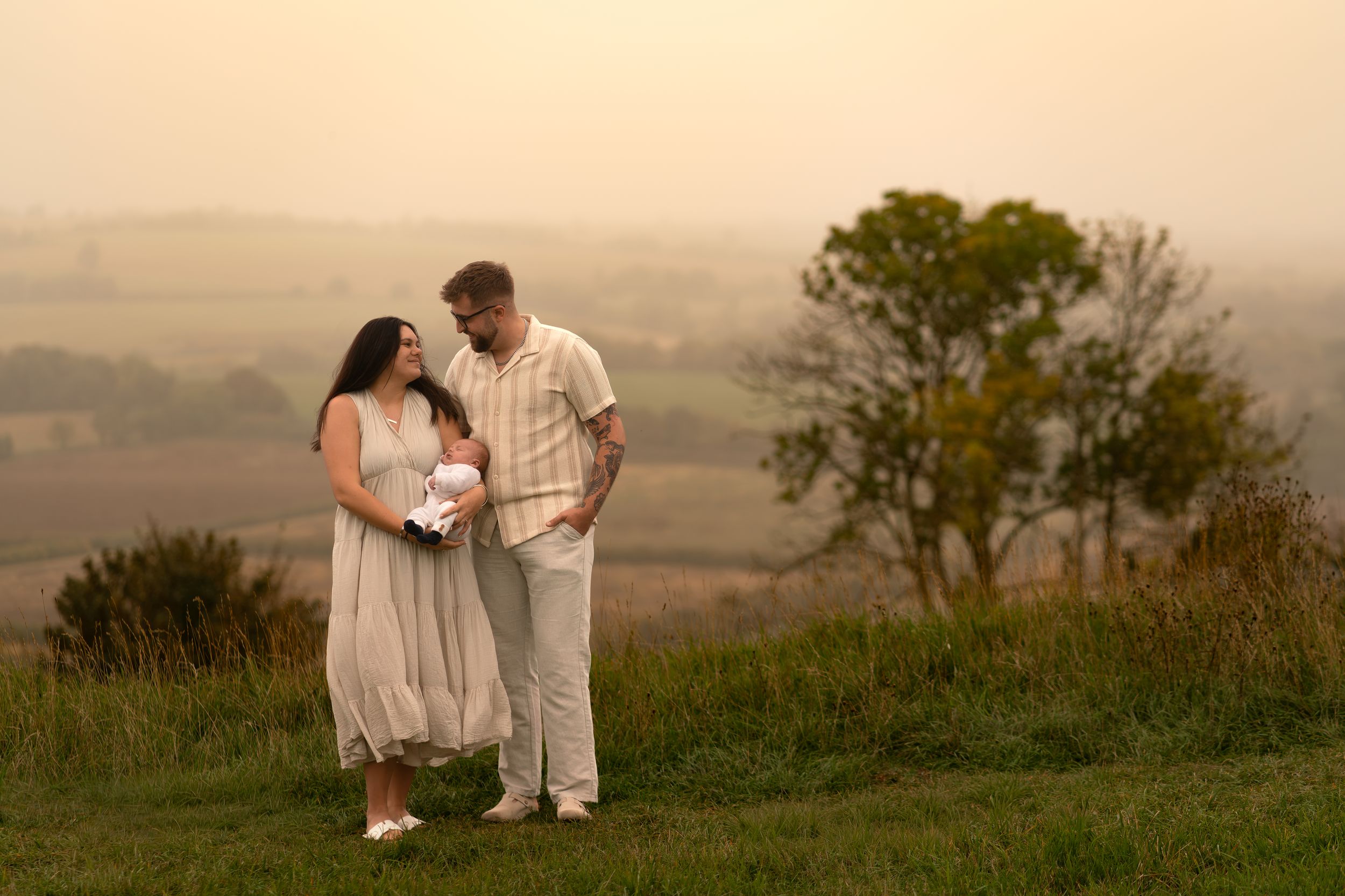Couple stands on a grassy hill at sunset, sharing a tender moment with a newborn baby in a misty countryside.