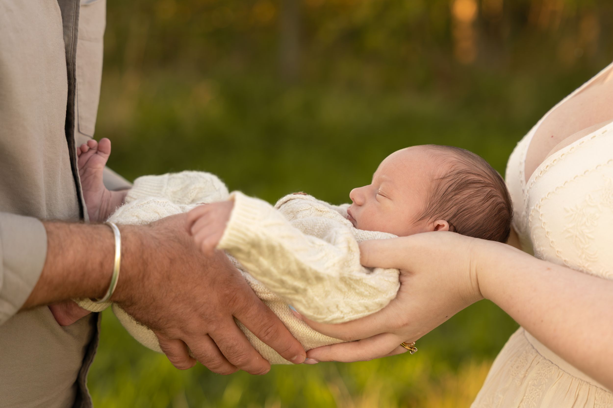 Parents cradling a sleeping newborn baby wrapped in white cloth outdoors with green bokeh background.
