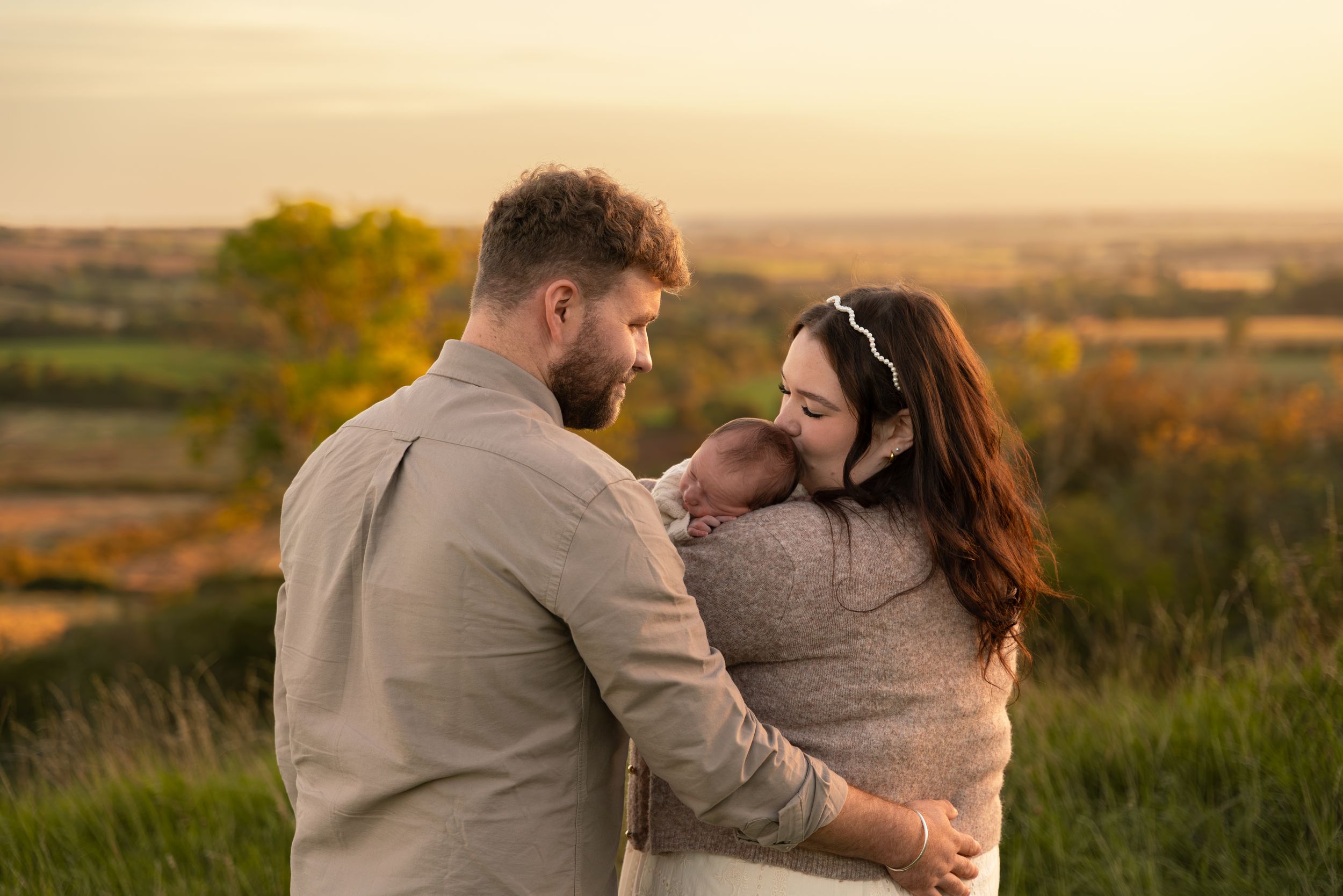 A couple shares a tender moment with their newborn baby at golden hour outdoors.