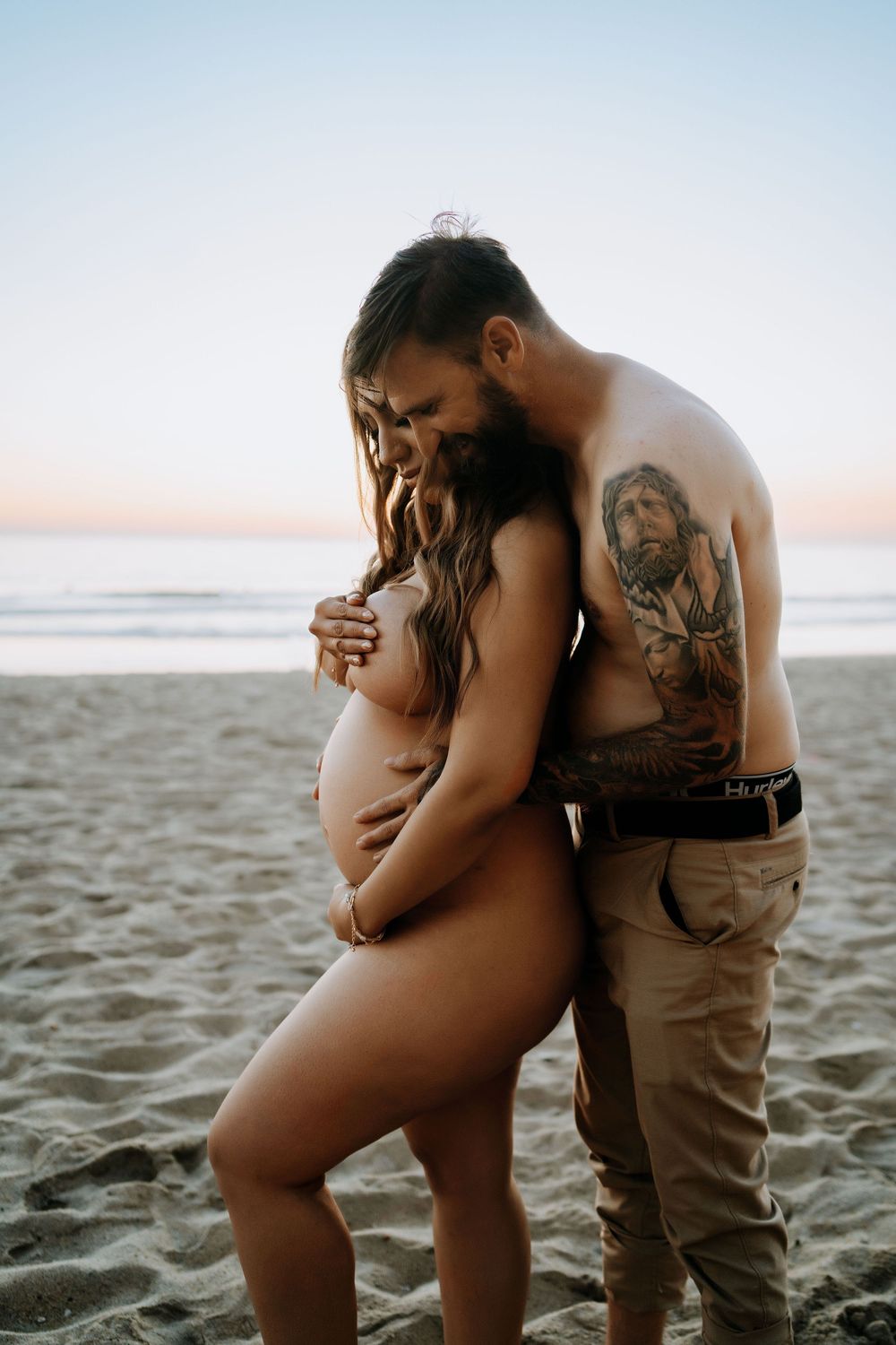 Pregnant couple embracing on sandy beach at sunset, man with tattoos holding woman from behind.
