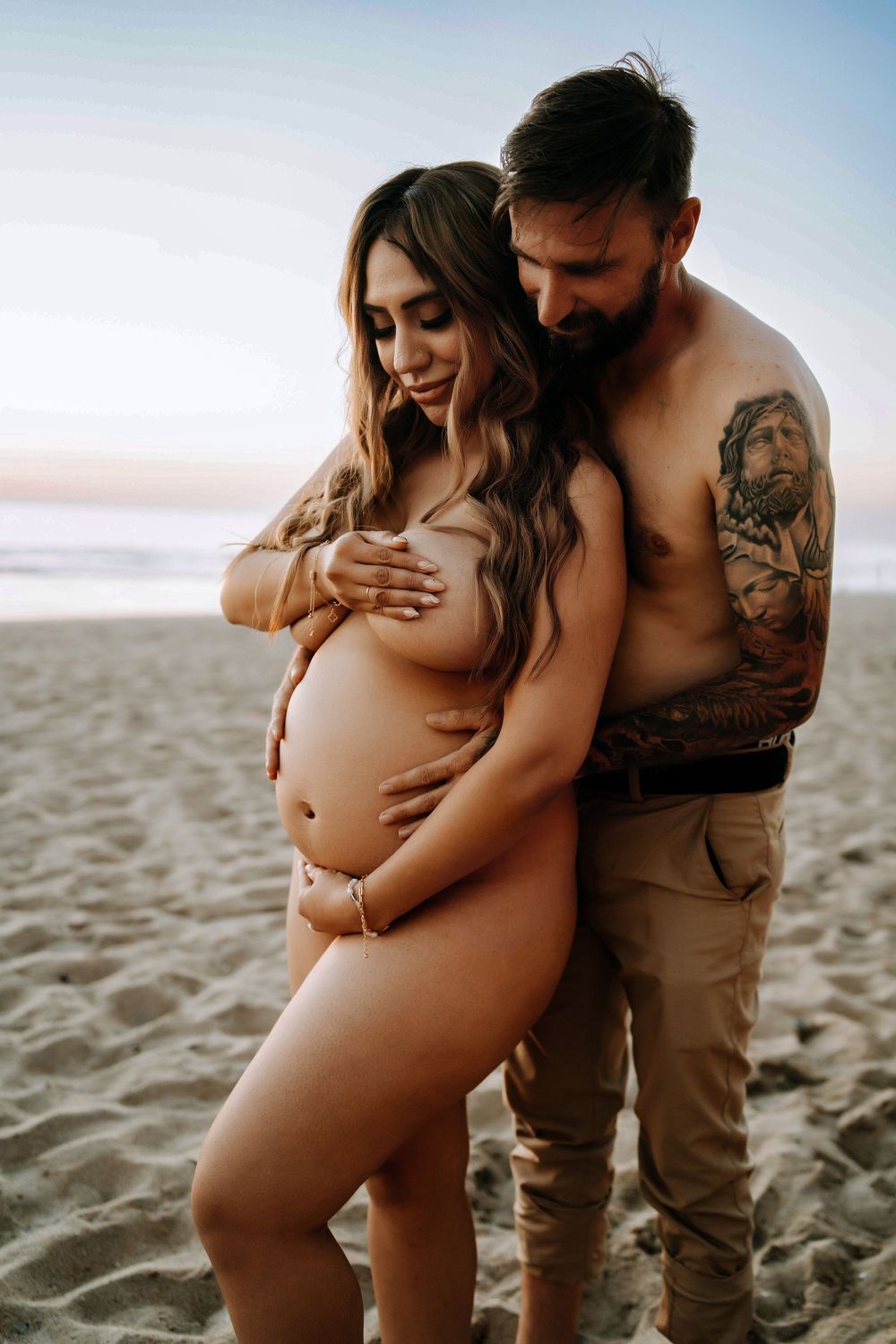Couple sharing intimate maternity beach photo at sunset, man with tattoos embracing pregnant woman on sandy shore.