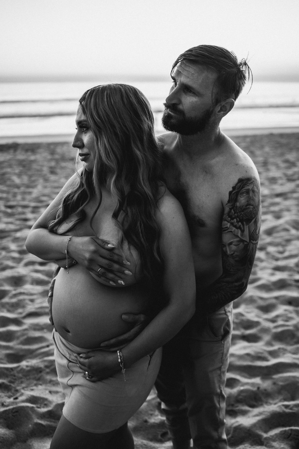 Black and white beach maternity photo of pregnant woman and tattooed shirtless man embracing on sandy shore.