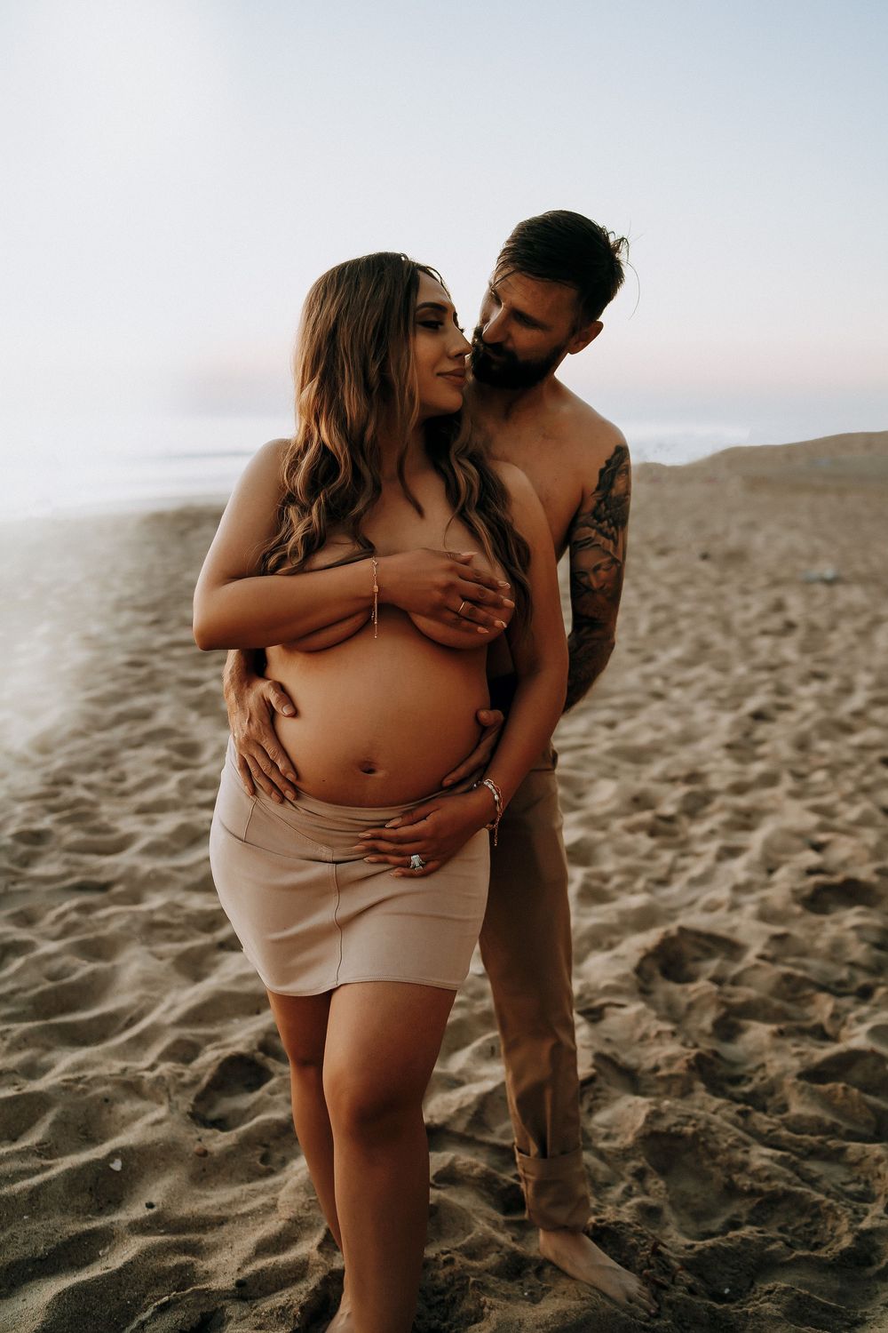 Couple sharing intimate maternity moment on sandy beach at dusk, man embracing pregnant woman from behind.