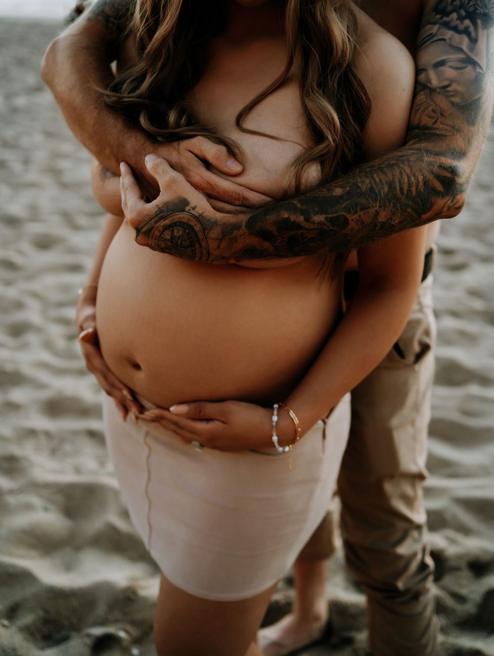 Pregnant woman on beach cradled by tattooed partner, both holding baby bump, maternity photo shoot.