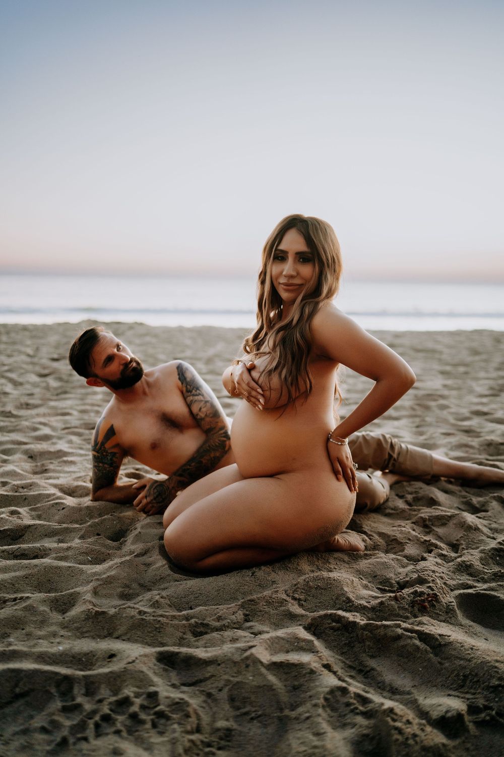 Pregnant woman and tattooed man sitting together on sandy beach at sunset in an artistic maternity photo.