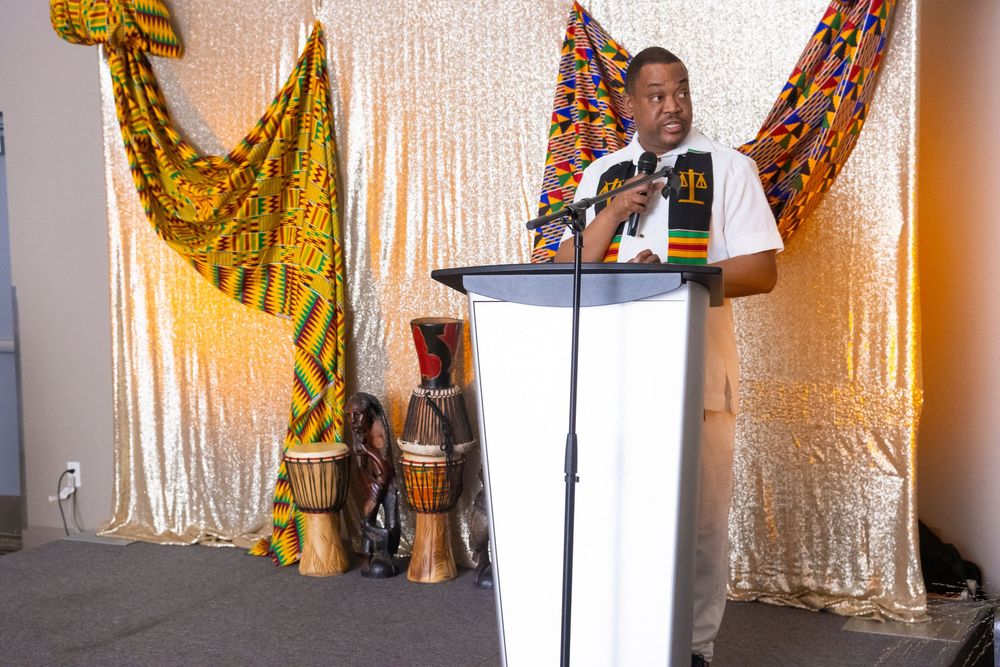 Man speaking at podium with African kente cloth backdrop and djembe drums at cultural event.