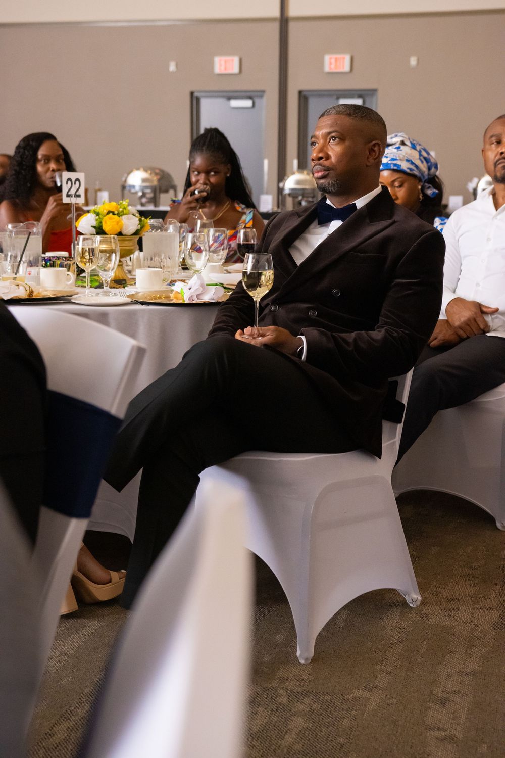 Man in black tuxedo holding wine glass seated at formal banquet event with guests at decorated tables.
