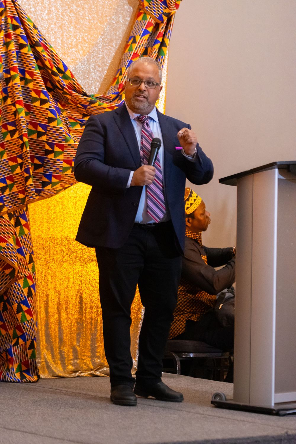Man in suit holding microphone speaks at podium before colorful African kente cloth backdrop at cultural event.