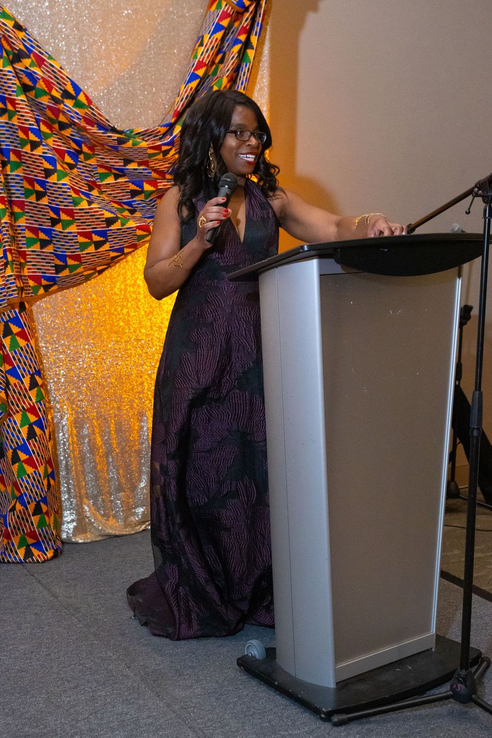 Woman in purple gown speaks at podium with microphone, colorful kente cloth backdrop at formal event.