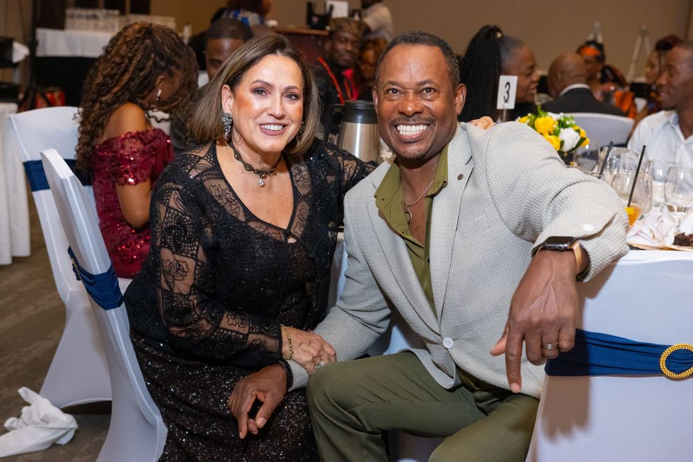 Two elegantly dressed guests smile and pose together at a formal banquet event with decorated tables in the background.