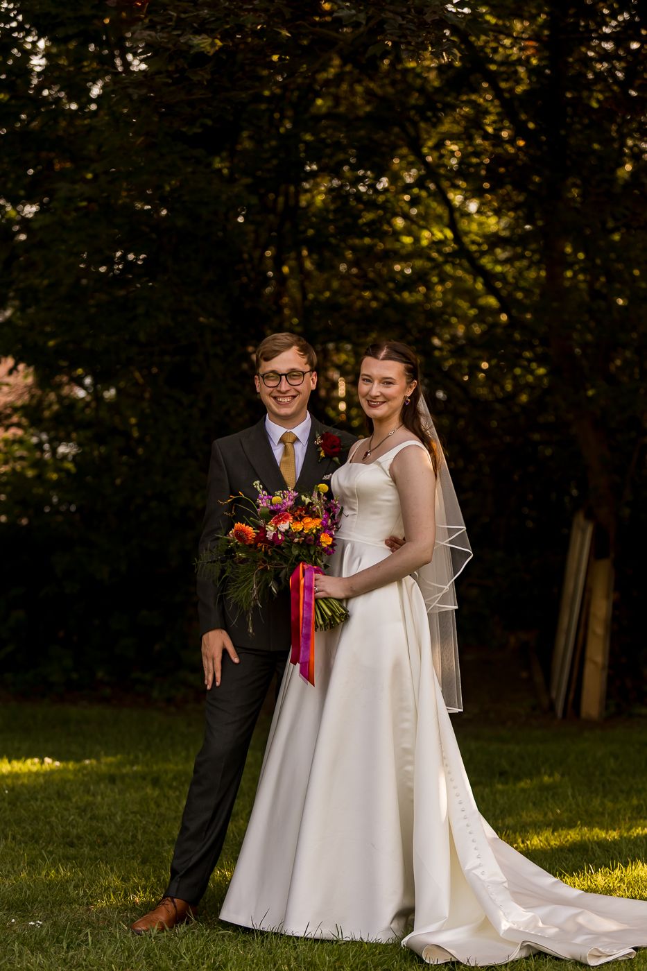 Bride and groom pose together outdoors holding a colorful bouquet, bride in white gown with veil, groom in dark suit.
