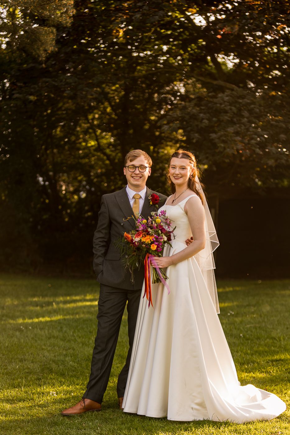 A smiling bride and groom pose together outdoors in golden hour light, she holds a colorful bouquet.