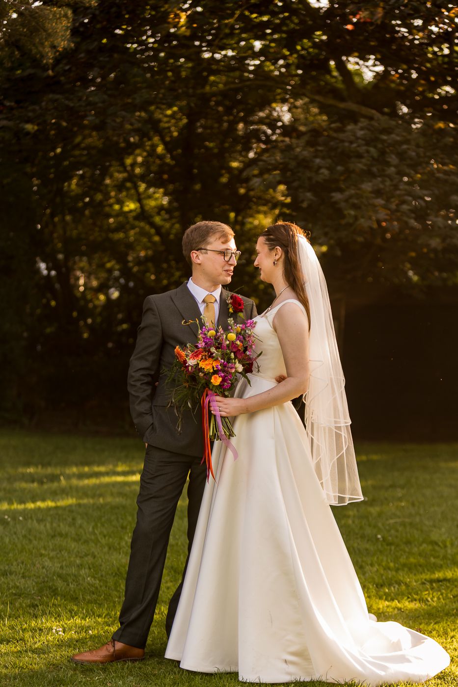 Bride and groom share a romantic moment outdoors, she holds a vibrant bouquet, golden sunlight filters through lush green trees.