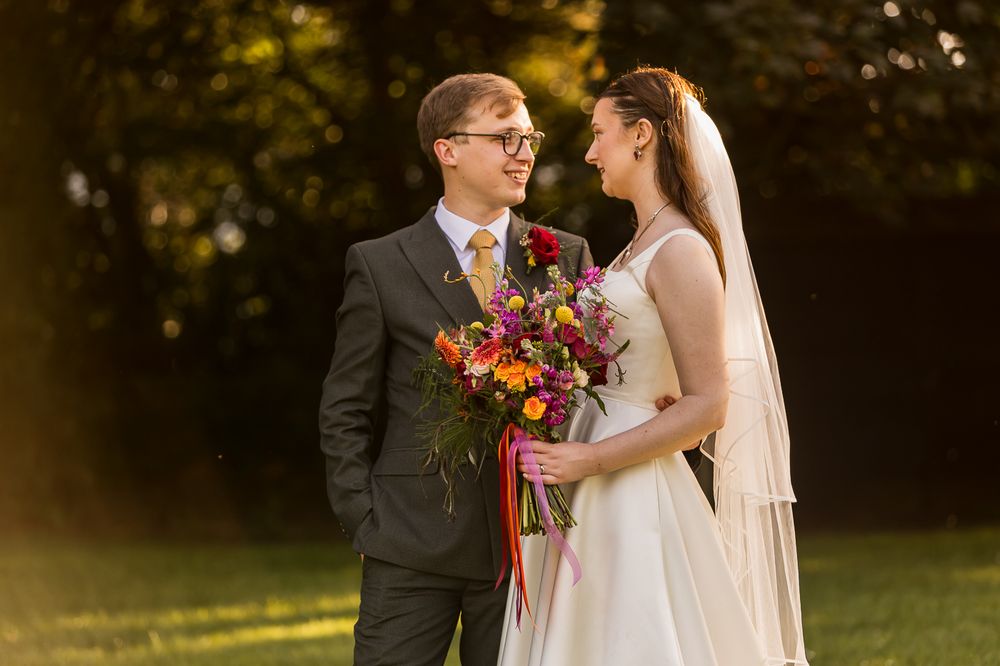 Bride and groom share a romantic moment outdoors, bride holding vibrant bouquet, golden sunlight glowing behind them.