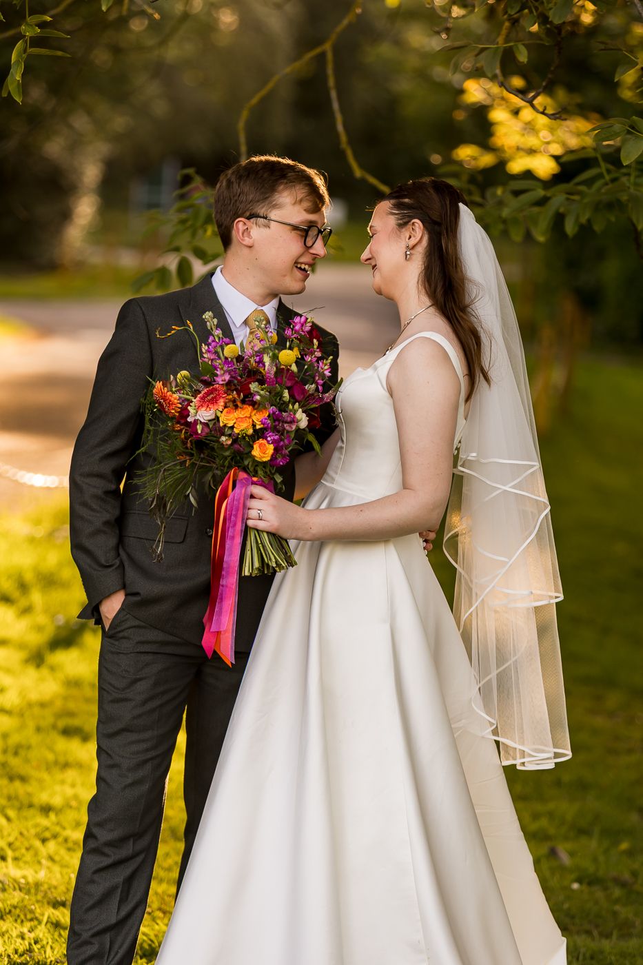 Smiling bride and groom stand close together under tree branches, sharing a joyful moment with colorful bridal bouquet.