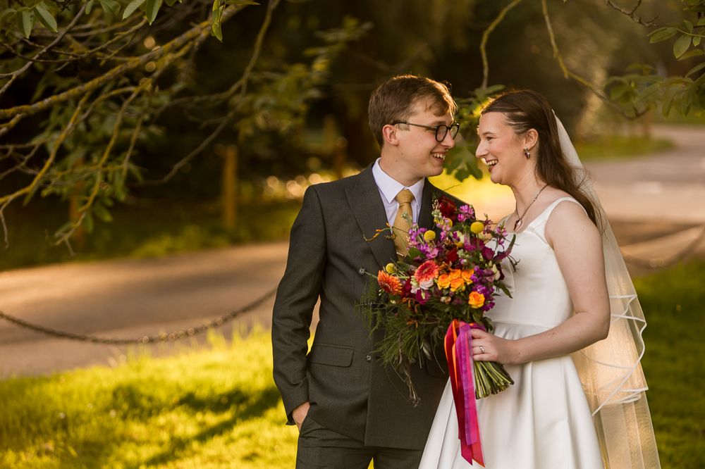 Bride and groom smiling at each other outdoors, holding a vibrant bouquet of colorful flowers in warm golden sunlight.