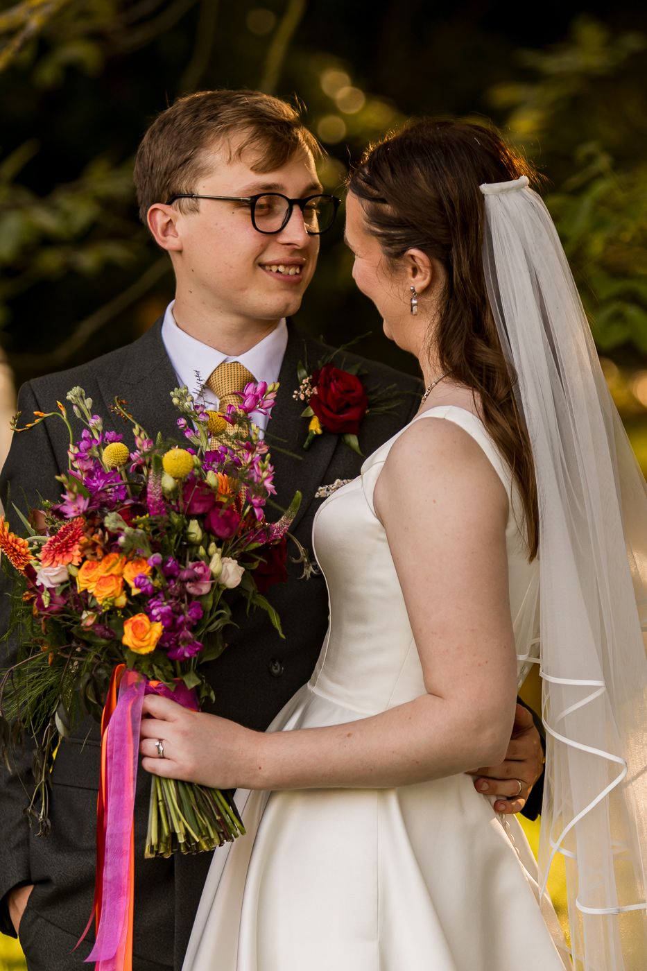 A bride in a white gown and veil holds a colorful bouquet while gazing at her groom in a suit with glasses.