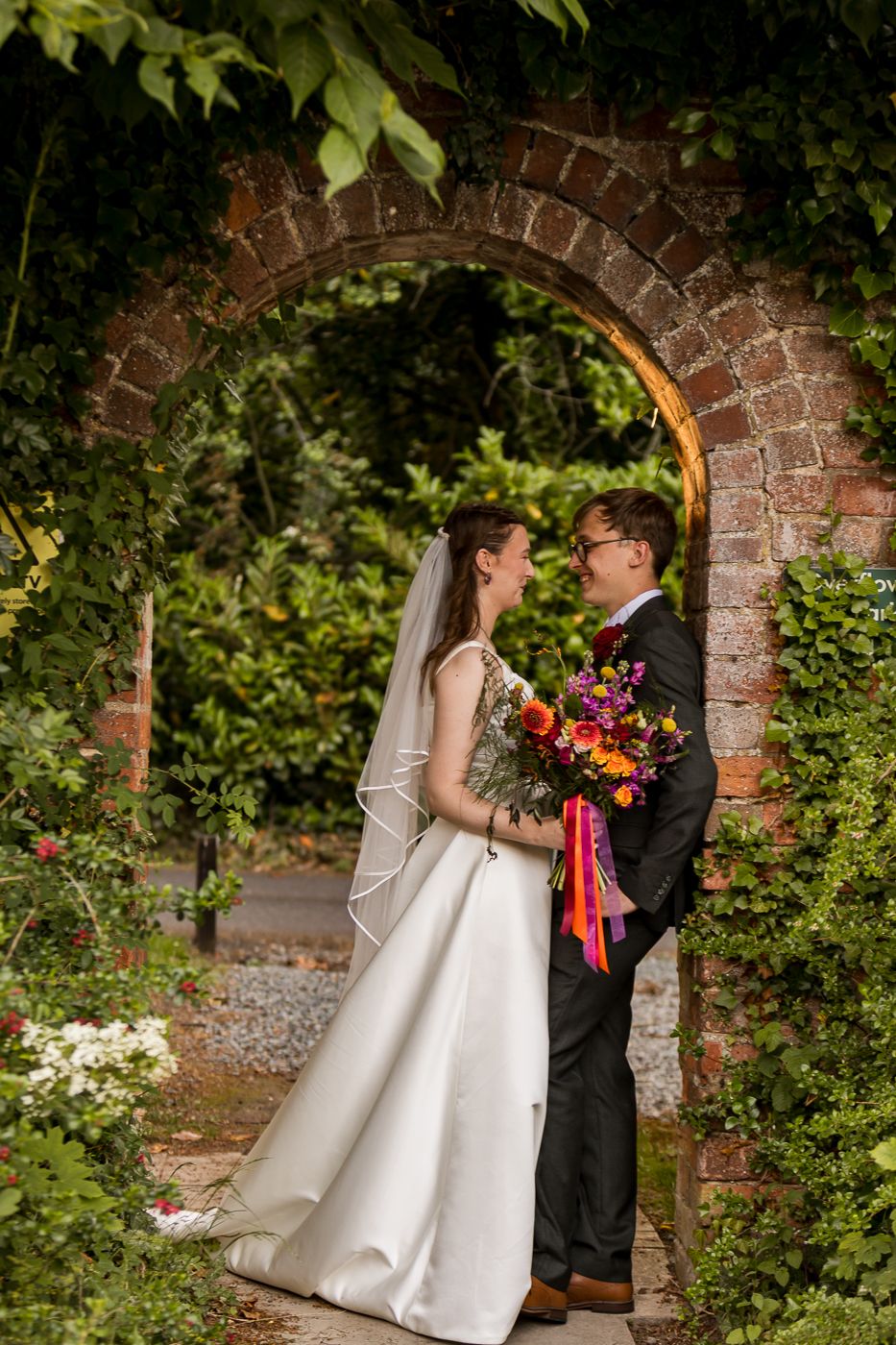 Bride and groom share a romantic moment under a brick arch in a lush garden, bride holding a vibrant bouquet.