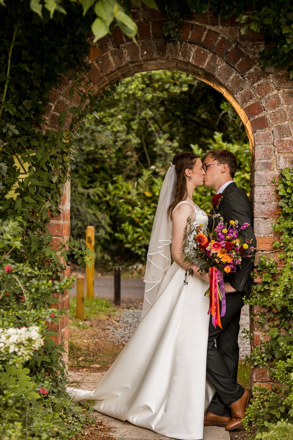 Bride and groom kissing under a rustic brick archway, bride holding colorful bouquet in a lush garden setting.