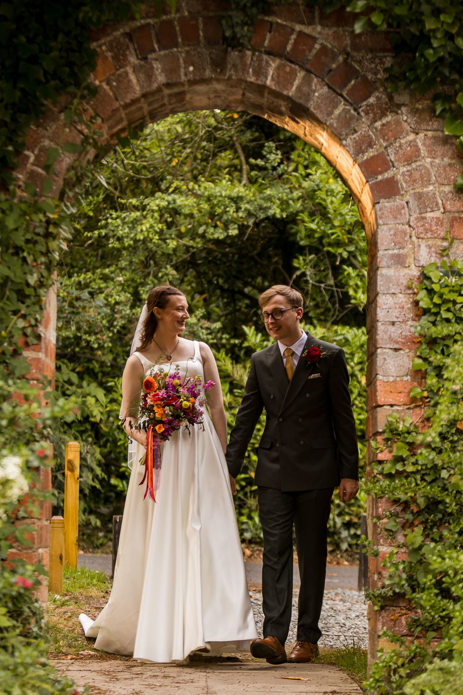 Bride and groom walking through a brick archway surrounded by lush greenery, bride holding a vibrant bouquet.