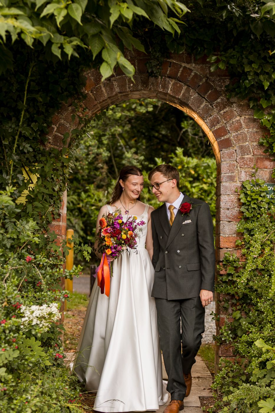 Bride and groom walk through brick garden arch, she holds colorful bouquet, both smiling in lush green setting.