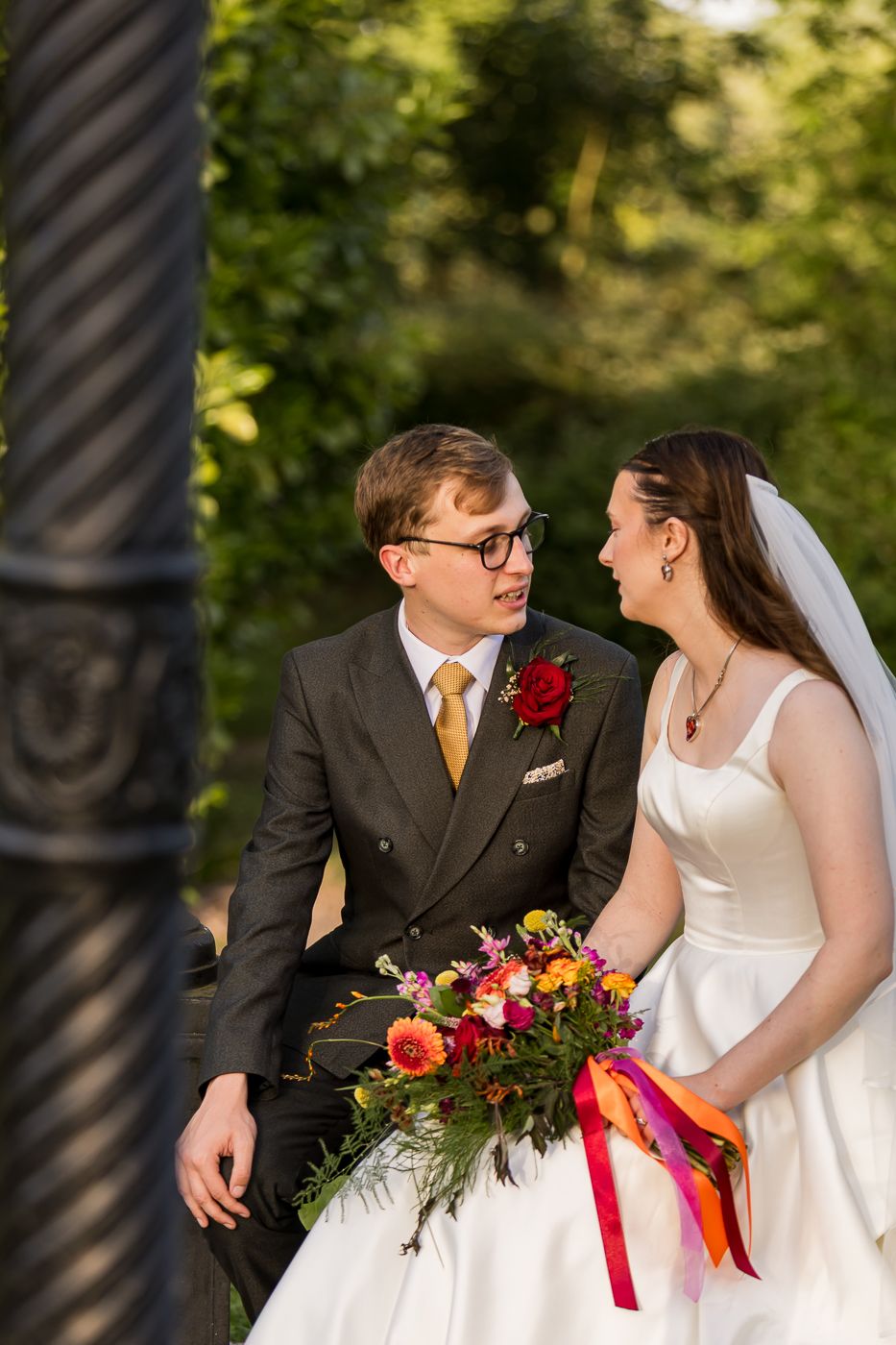 Bride and groom share a tender moment outdoors, she holds a vibrant bouquet with colorful ribbons, he wears a dark suit with gold tie.