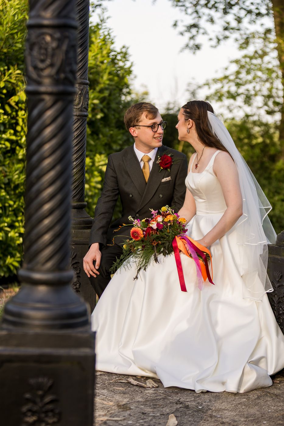 A bride and groom share a romantic moment outdoors, she holding a colorful bouquet with ribbons, both dressed in wedding attire.