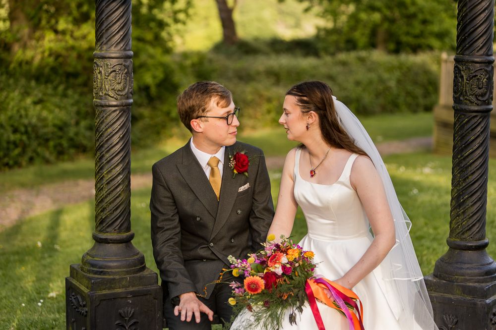 Bride and groom sitting together under ornate iron gazebo, sharing a loving glance, with colorful bouquet in lush green park.