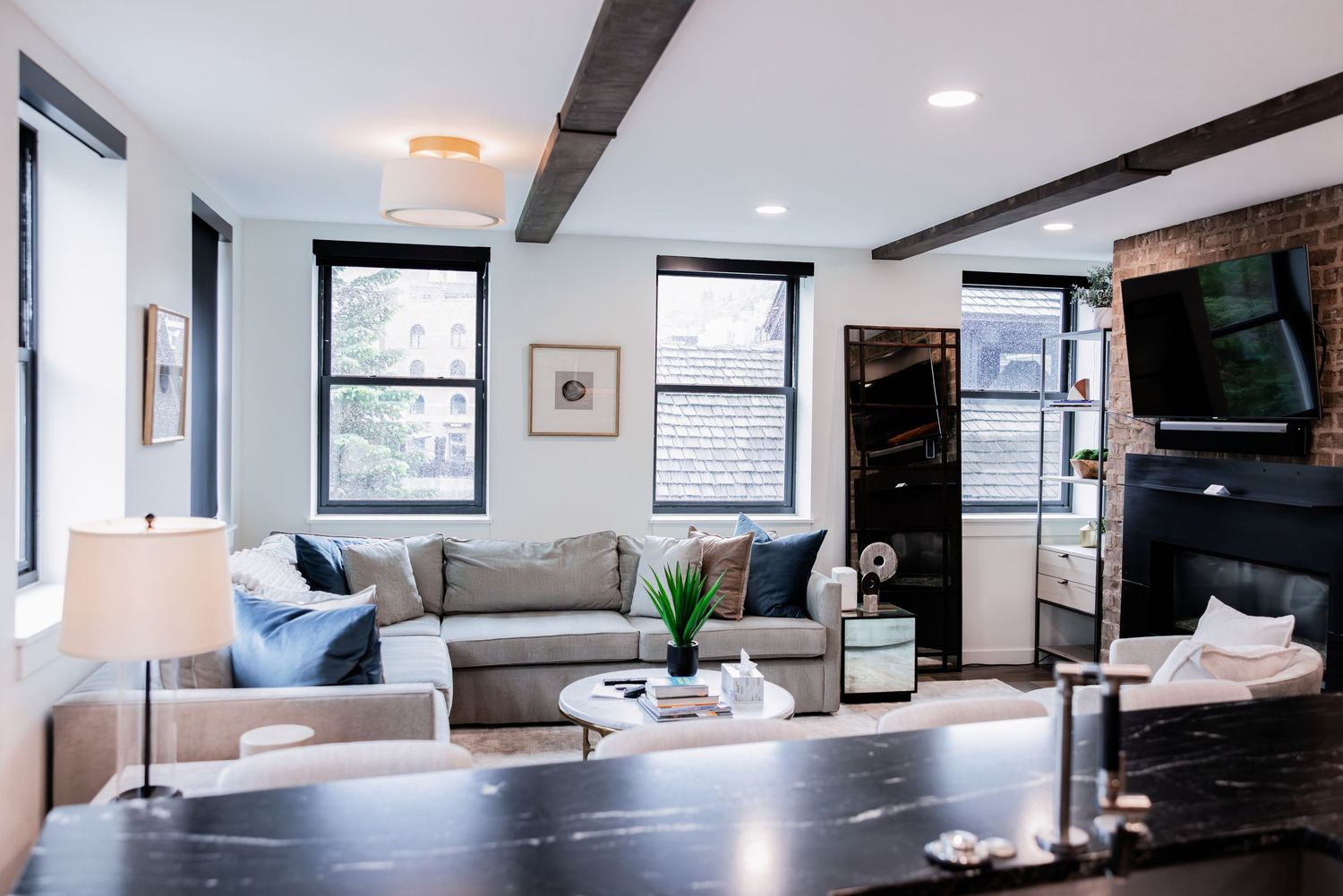 Bright modern living room with gray sectional sofa, exposed wooden ceiling beams, large windows, wall-mounted TV and a dark granite counter.