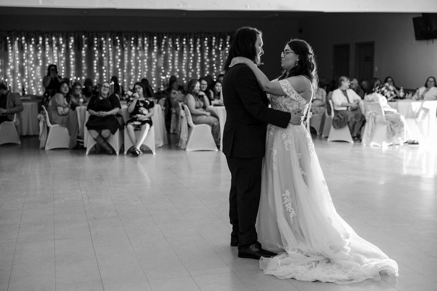 Black and white photo of a bride and groom sharing their first dance at a wedding reception with guests seated in background.