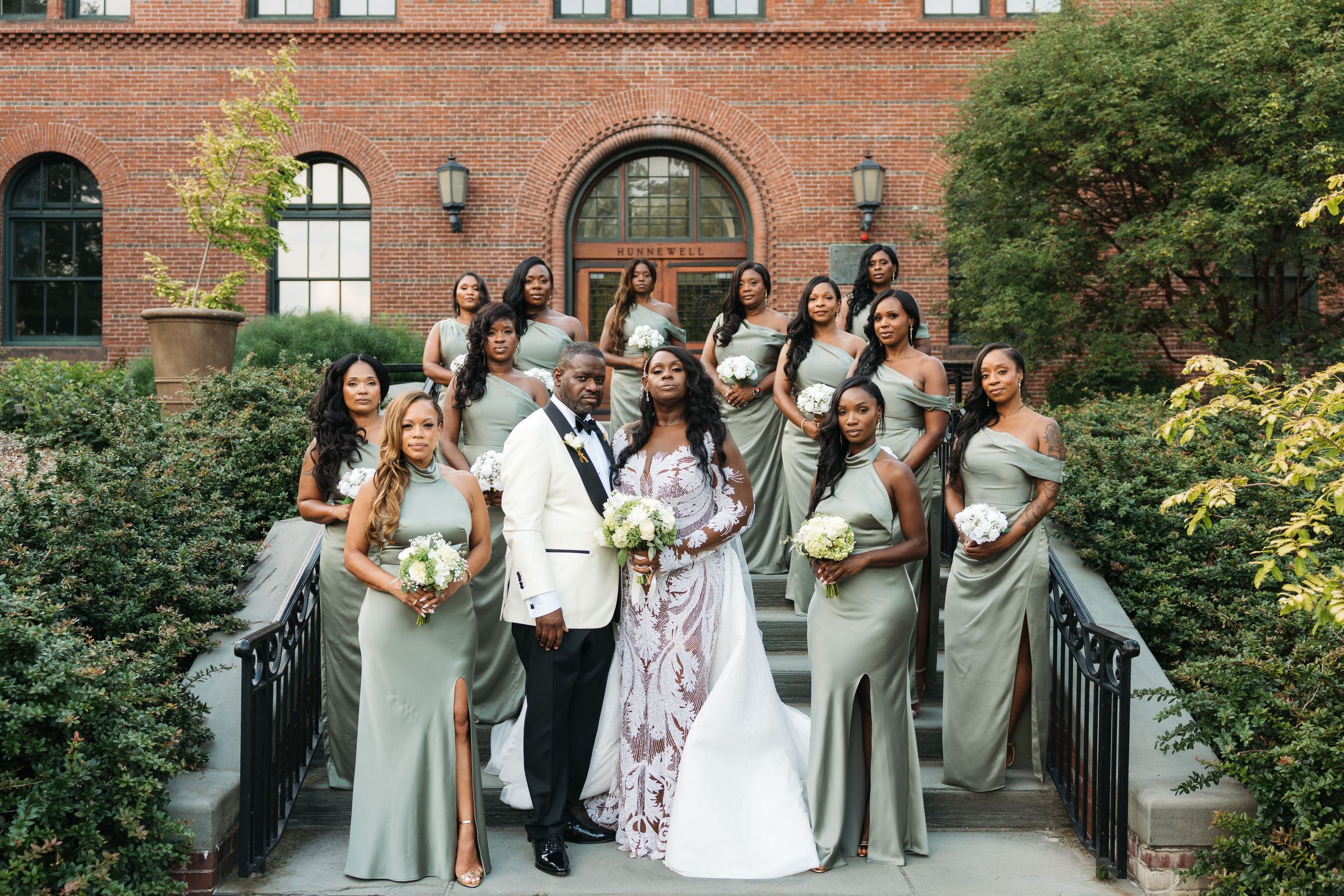 Bride and groom pose with bridesmaids in sage green gowns on brick building steps at elegant outdoor wedding.