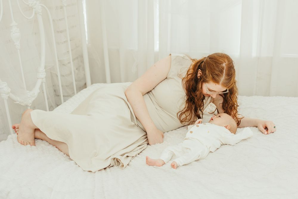 A red-haired mother in a cream dress tenderly kisses her newborn baby on a white bed.
