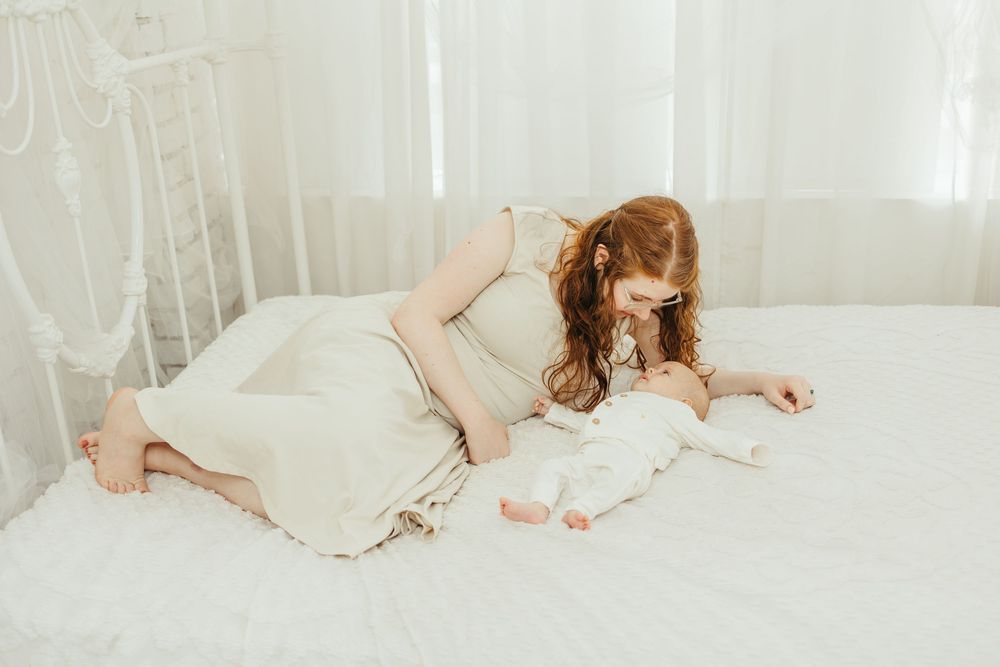 A mother in a cream dress lies on a white bed, gazing lovingly at her newborn baby in a bright, airy room.