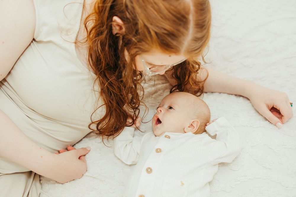 Red-haired mother gazing lovingly at her yawning newborn baby dressed in white on a soft blanket.