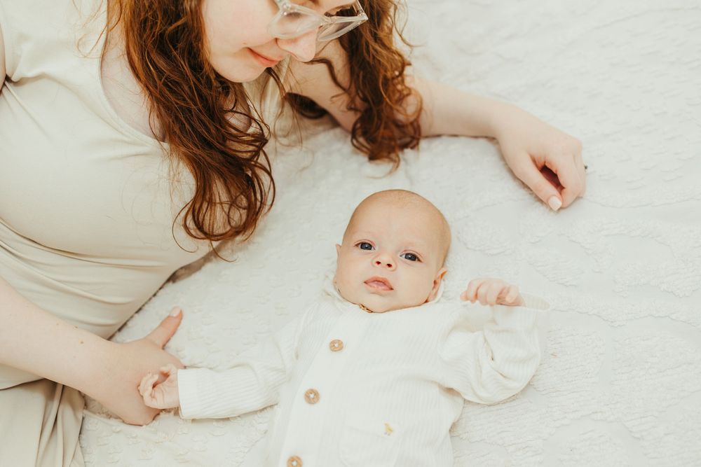 A red-haired mother lying on a white blanket smiling at her newborn baby in a white button-up onesie.
