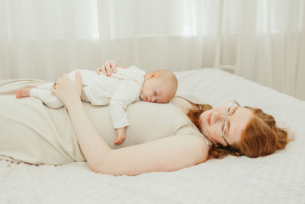A mother with red hair lies on a white bed smiling, cradling her sleeping newborn baby on her chest.