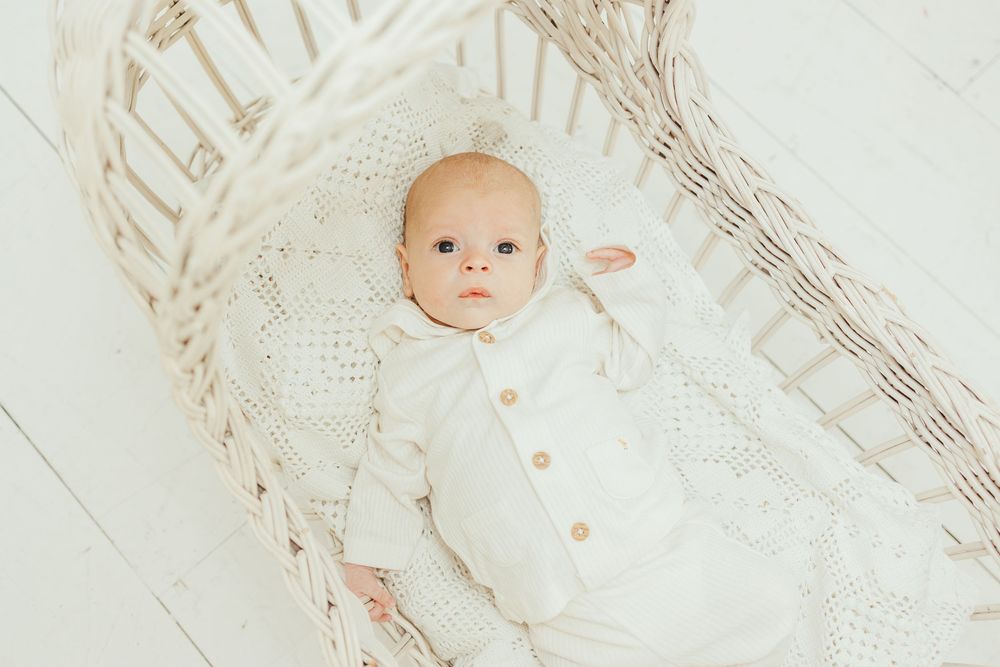 A newborn baby in a white outfit lying in a wicker bassinet with soft white bedding.
