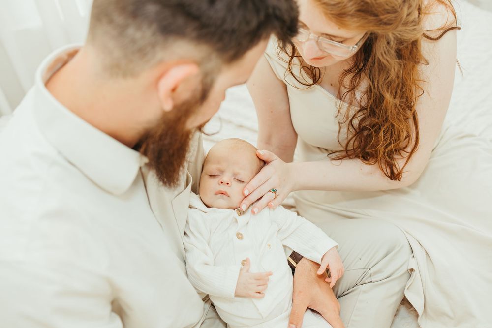 A bearded father and redheaded mother tenderly look at their newborn baby dressed in white on a bed.