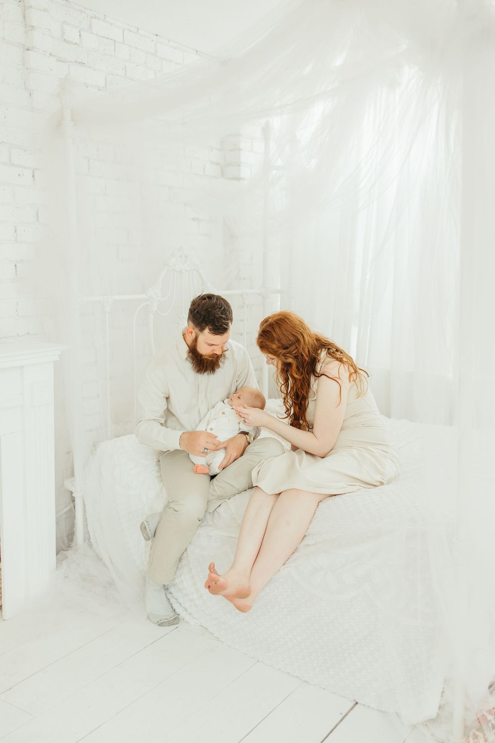 A bearded man and redhead woman in white outfits sit on a bed holding their newborn baby in a bright white room.